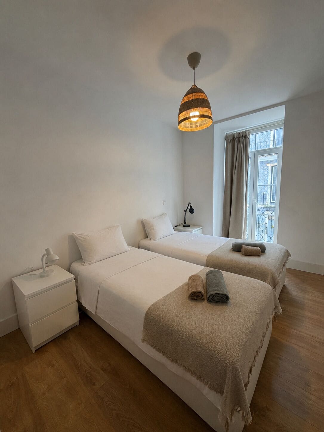 Ein Hotelzimmer mit zwei Betten, einem Nachttisch mit Lampe, einer Pendelleuchte und einem Fenster mit Vorhängen.
