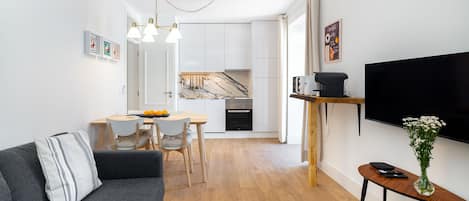 En moderne stue med en grå sofa, et spiseområde med et træbord og stole, et køkken med hvide skabe og et marmorbeslag, samt et ophængt tv på et træstel.