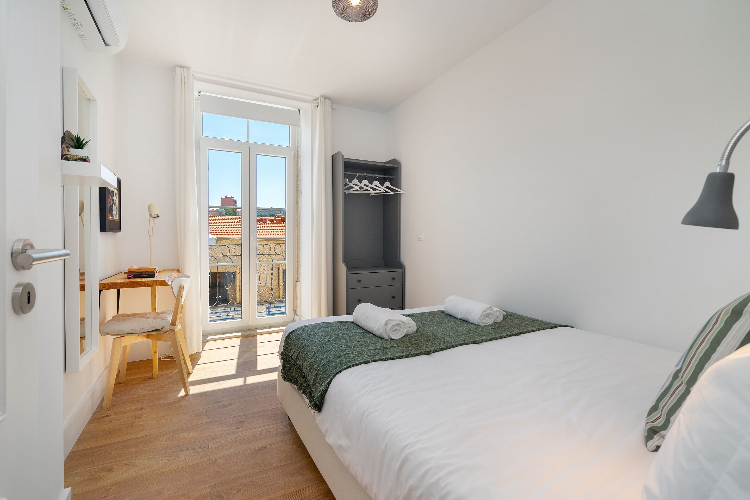 Un dormitorio con cama, escritorio, silla, armario y ventana con vista a edificios.