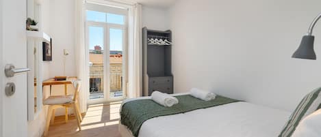 Un dormitorio con cama, escritorio, silla, armario y ventana con vista a edificios.