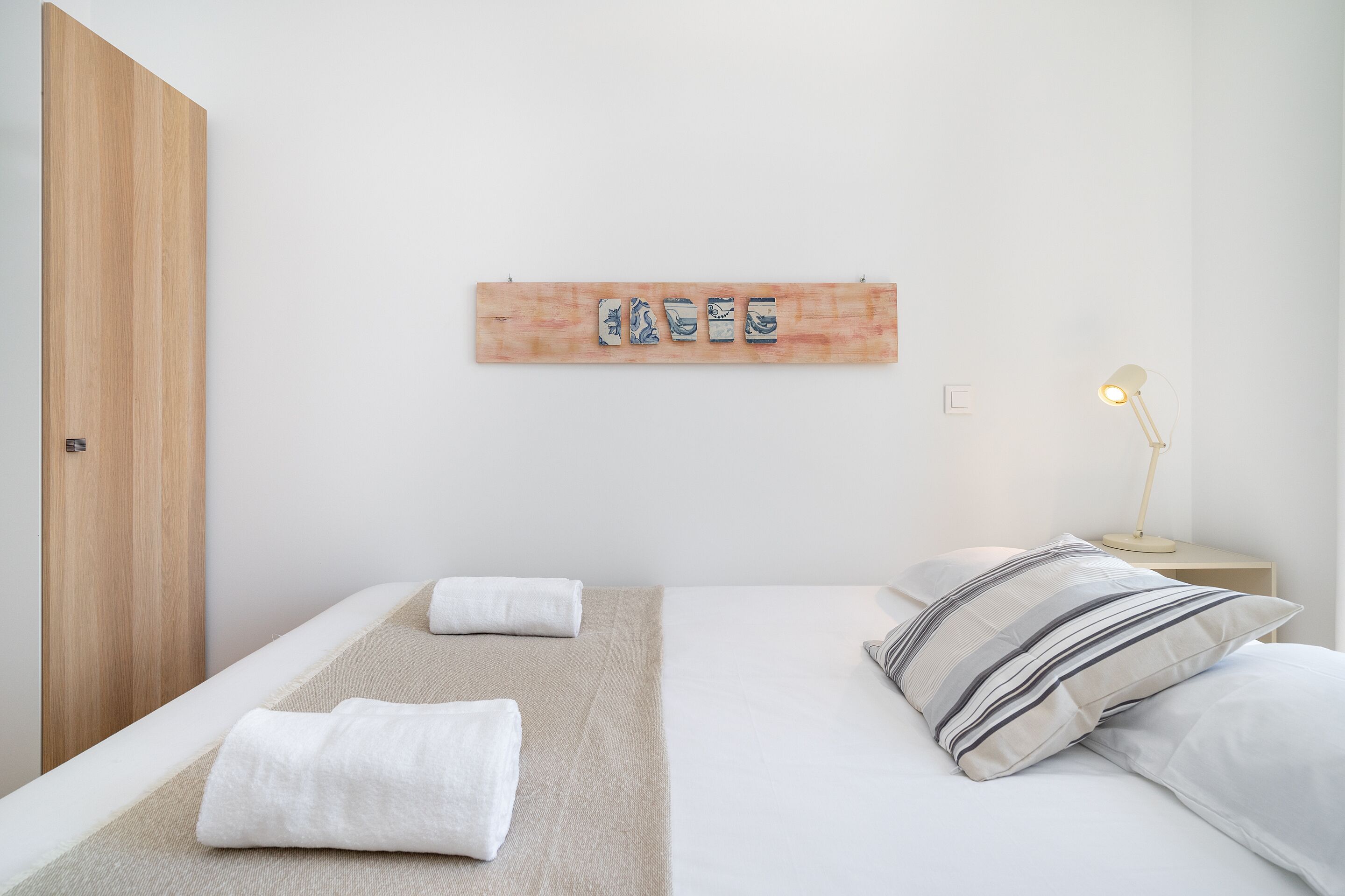Quarto de hotel minimalista com uma cama, duas almofadas, uma luminária de cabeceira e uma obra de arte na parede.