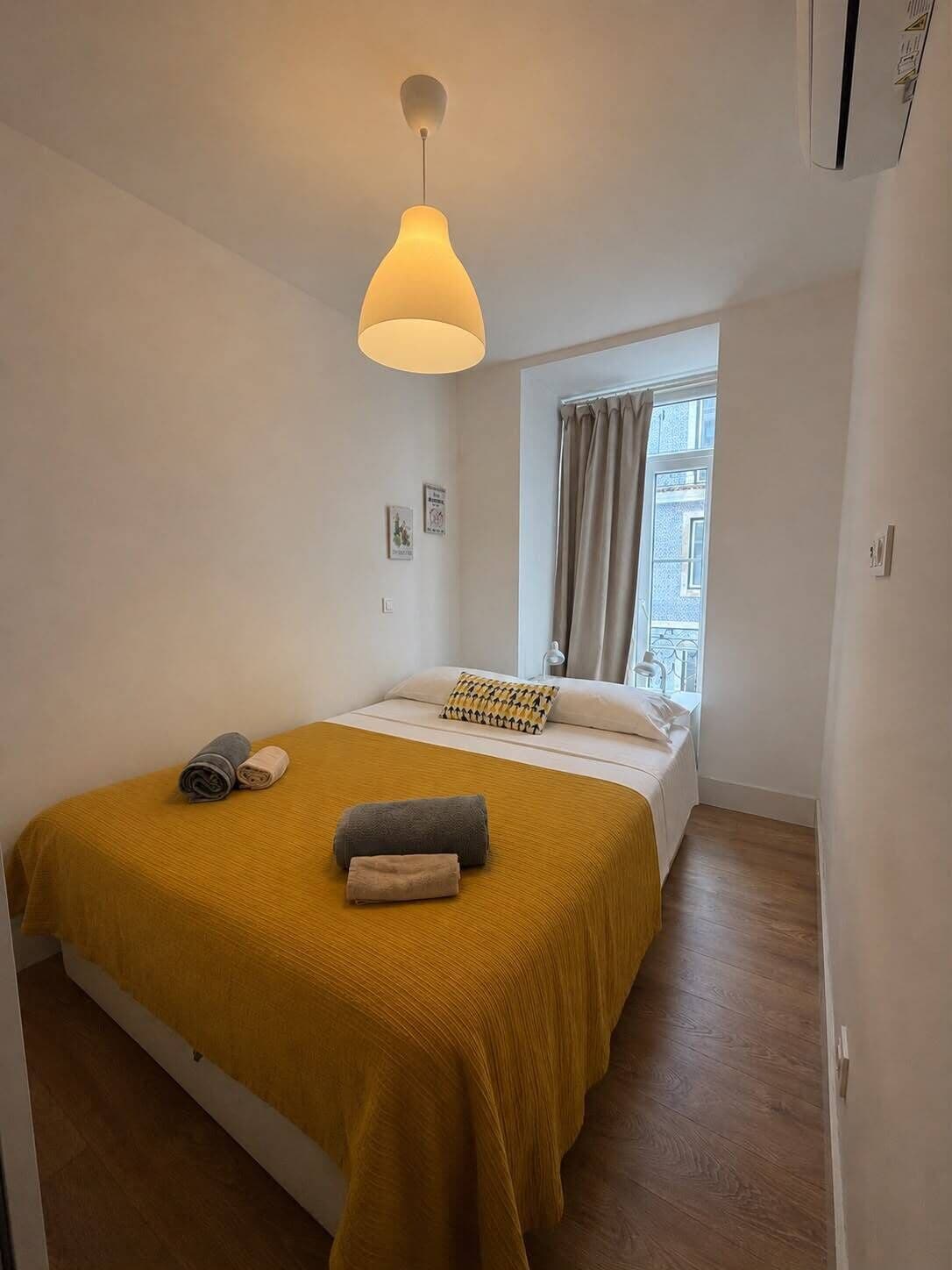 A hotel room with a bed, a window with curtains, a hanging light fixture, and a wall-mounted air conditioning unit.