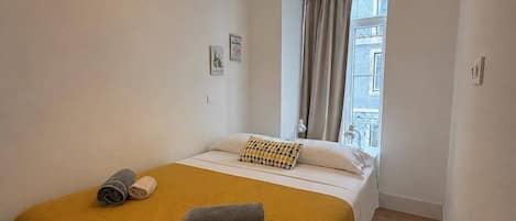 A hotel room with a bed, a window with curtains, a hanging light fixture, and a wall-mounted air conditioning unit.