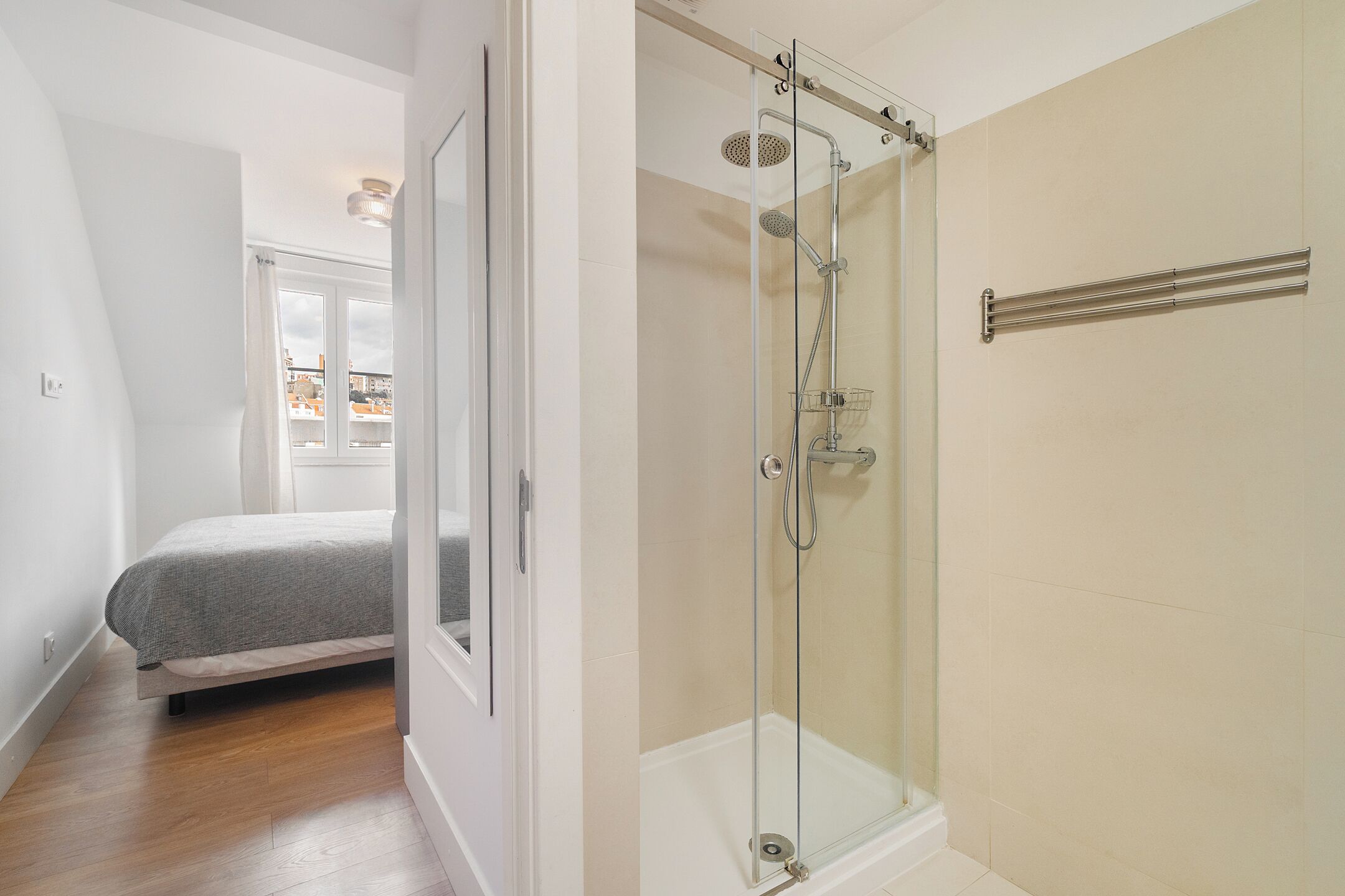A modern bathroom with a glass shower enclosure, a visible towel rack, and a bed in the adjacent room.