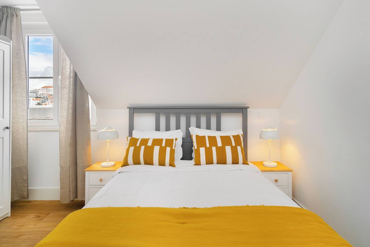 A bedroom with a bed, two nightstands, a window with curtains, and a view of buildings outside.