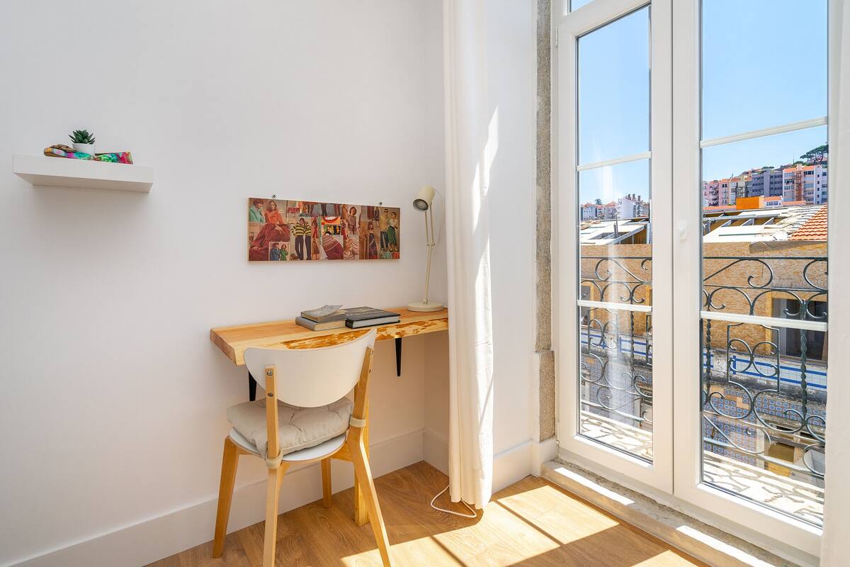 A room with a wooden desk, a white chair, a small shelf with a plant, and a large window with a view of buildings.