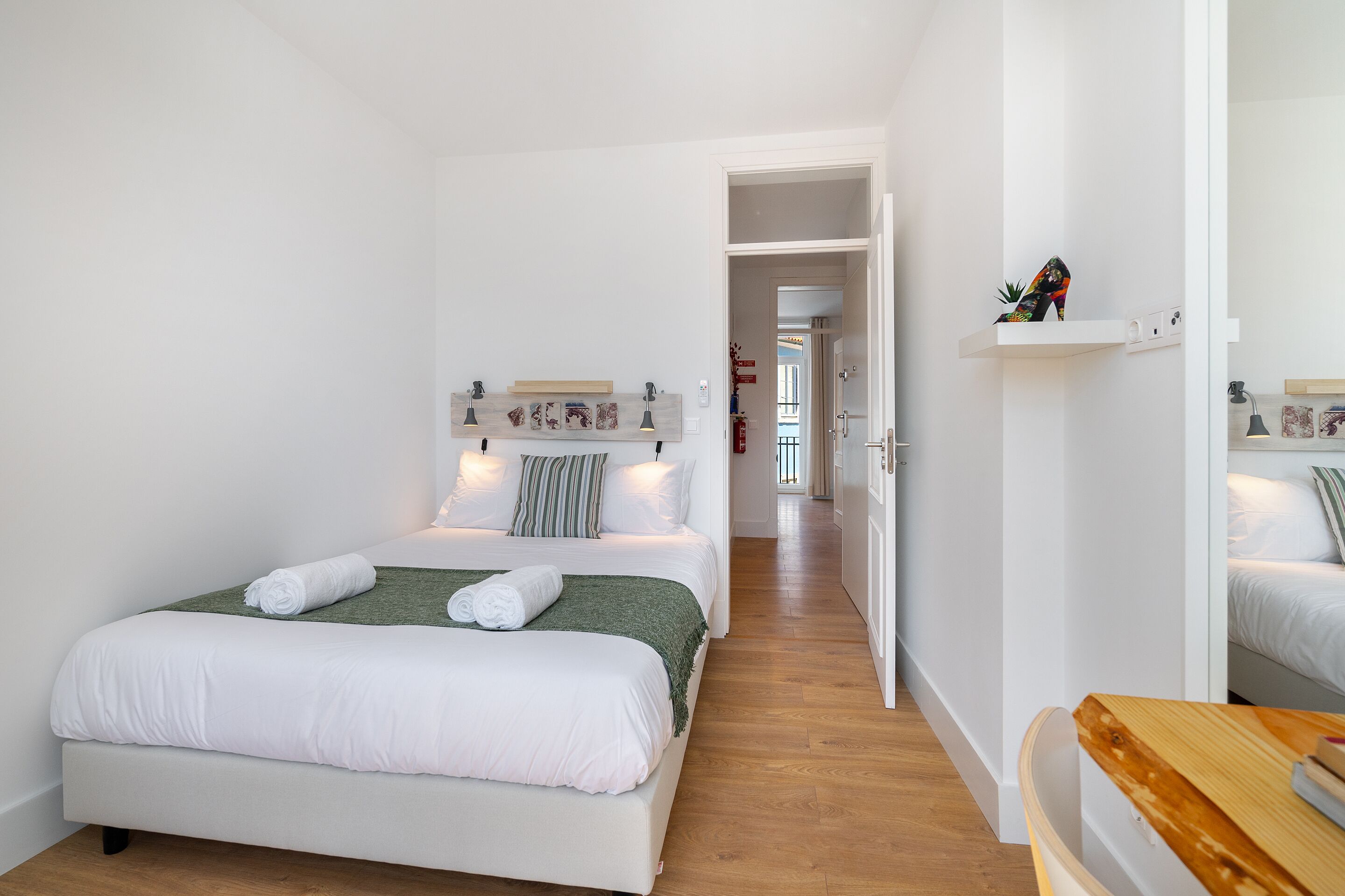 A modern hotel room with a large bed, a wooden bench, and a hallway leading to another room.