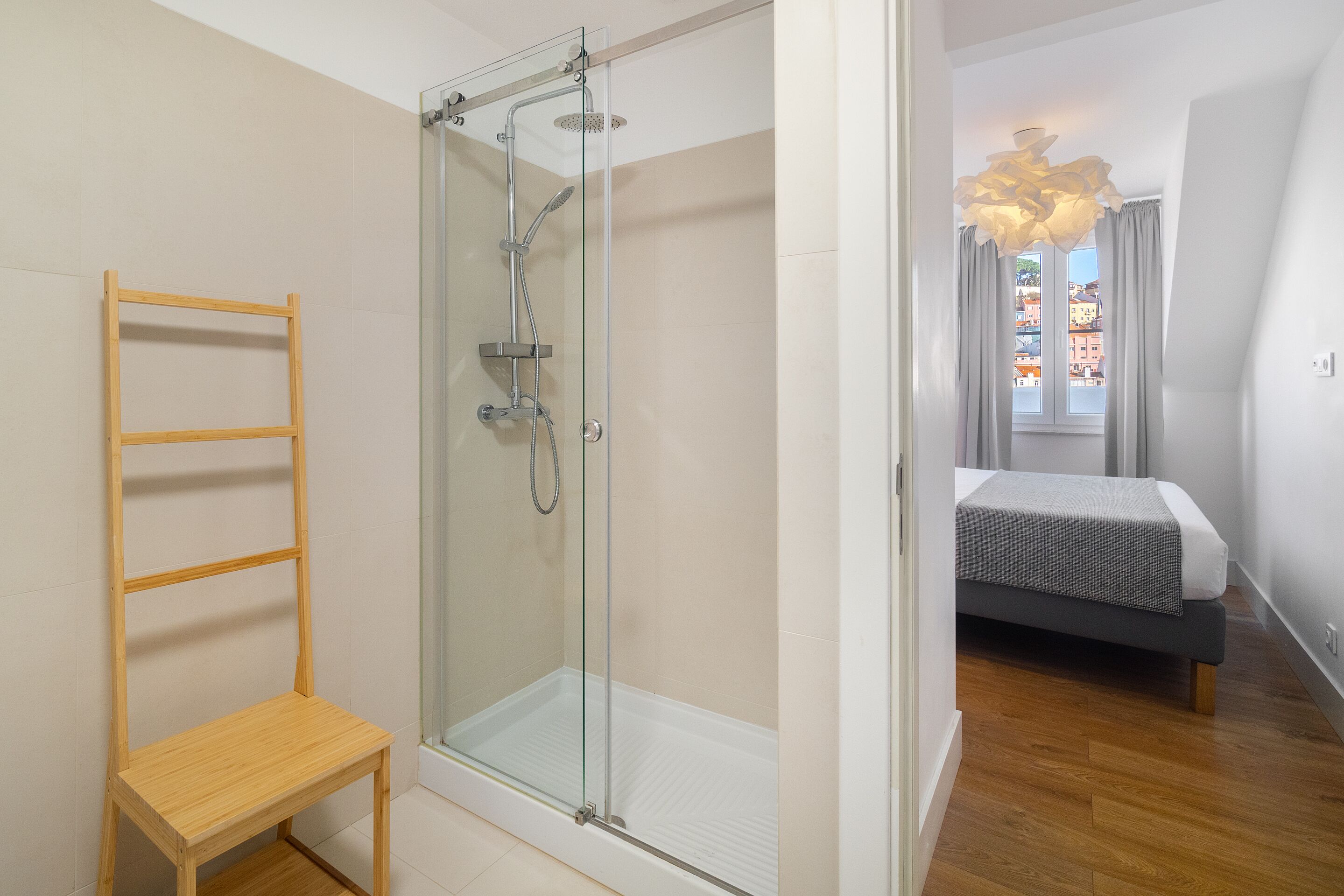 A modern bedroom with a glass shower enclosure, a wooden chair, and a bed with a grey and white bedding.