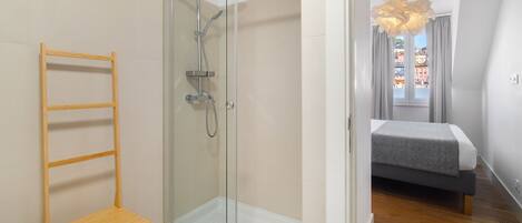 A modern bedroom with a glass shower enclosure, a wooden chair, and a bed with a grey and white bedding.