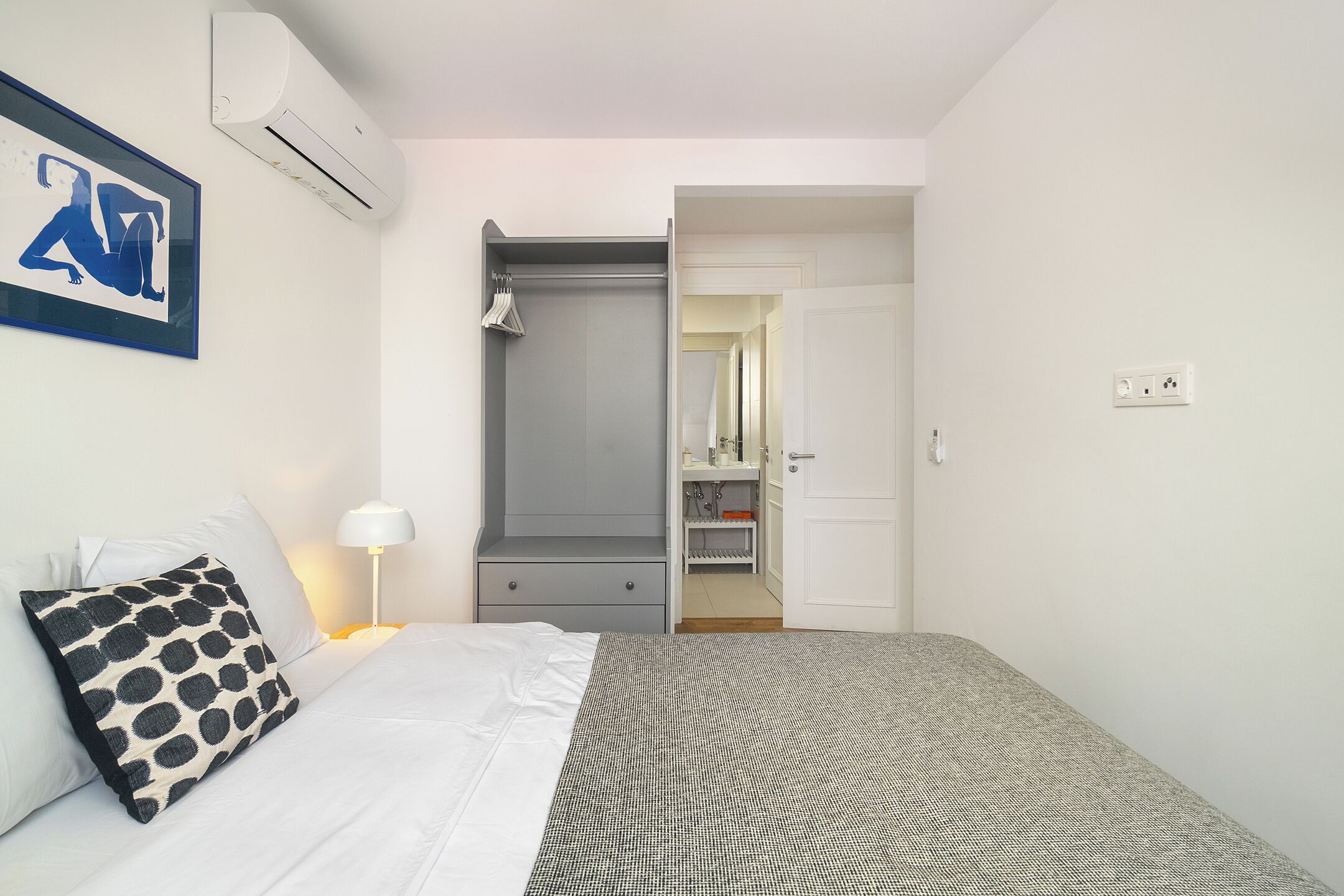 A hotel room with a bed, a gray patterned area rug, a white wall with a framed picture, a gray wardrobe, a white bedside table with a lamp, and a bathroom visible through an open door.