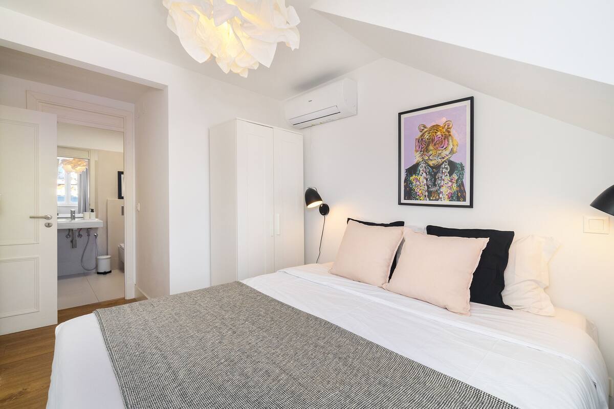 A bedroom with a large bed, a white wardrobe, a bathroom visible through an open door, and a wall-mounted air conditioner.