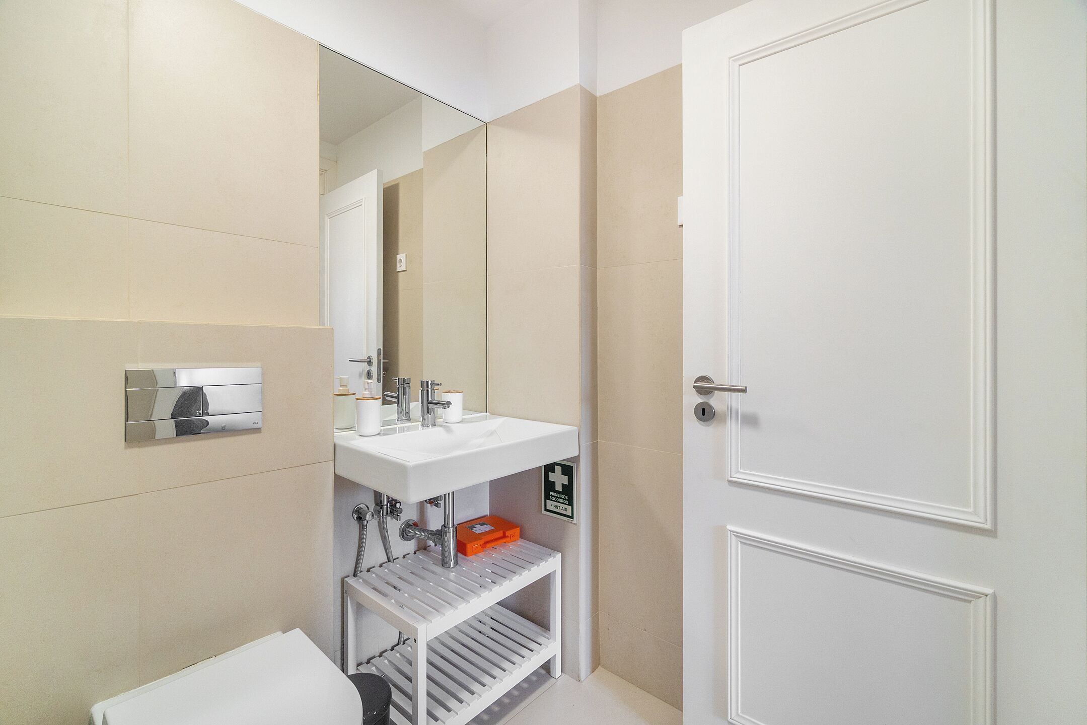 Une salle de bain moderne avec un lavabo blanc, un miroir et des toilettes. On y trouve une étagère avec des articles de toilette et une trousse de premiers secours.