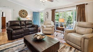 A living room with a brown leather sofa, beige armchairs, a wooden coffee table with decorative items, a ceiling fan, and a large window with curtains. - Twin Ponds Retreat-Pond/Hot Tub/Fire Pit/Game Room/Pet Friendly by RedAwning (Whittier)