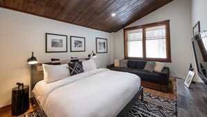 Een slaapkamer met een bed, een slaapbank, een raam met zonwering en ingekaderde foto's aan de muur.