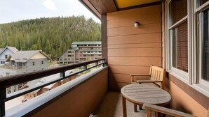 A balcony with a wooden table and chair, a window with blinds, and a view of a forested hillside and buildings. - Mountain View Ski-In Ski-Out Condo - Zephyr Mountain Lodge Premium-Rated 2516 (Winter Park)