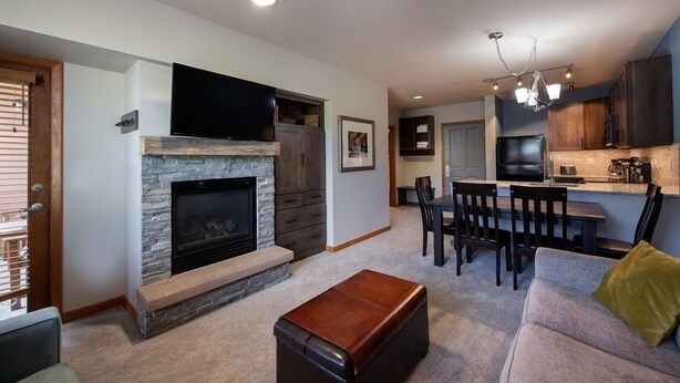 A modern living room with a stone fireplace, a flat-screen TV mounted above it, and a dining area with a kitchen in the background. - Mountain View Ski-In Ski-Out Condo - Zephyr Mountain Lodge Premium-Rated 2516 (Winter Park)