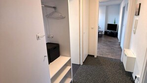 Een hal met tapijttegels, een kast met hangende kleding, een televisie in de aangrenzende kamer en een witte muur met een telefoon.