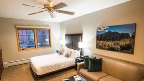 A hotel room with a bed, a sofa, a coffee table, a ceiling fan, a window with a view of trees, and a painting on the wall. - Grand Timber Lodge Studio Ski in/out (Breckenridge)