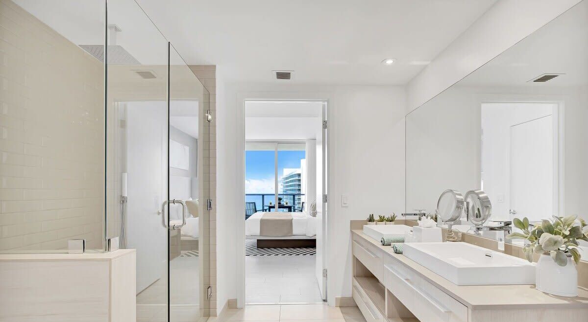 A modern bathroom with a glass shower enclosure, a large mirror, and a view of a bedroom with a bed and a cityscape.
