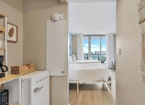 Habitación de hotel con cama, minibar, pequeño escritorio con cafetera y vistas a la ciudad a través de amplias ventanas.