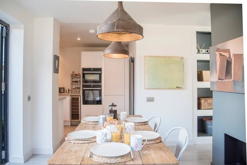 A dining area with a wooden table set for four, a hanging pendant light, and a kitchen in the background.