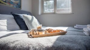 A neatly made bed with a tray of croissants and coffee, a window with a view of trees, and a painting on the wall.