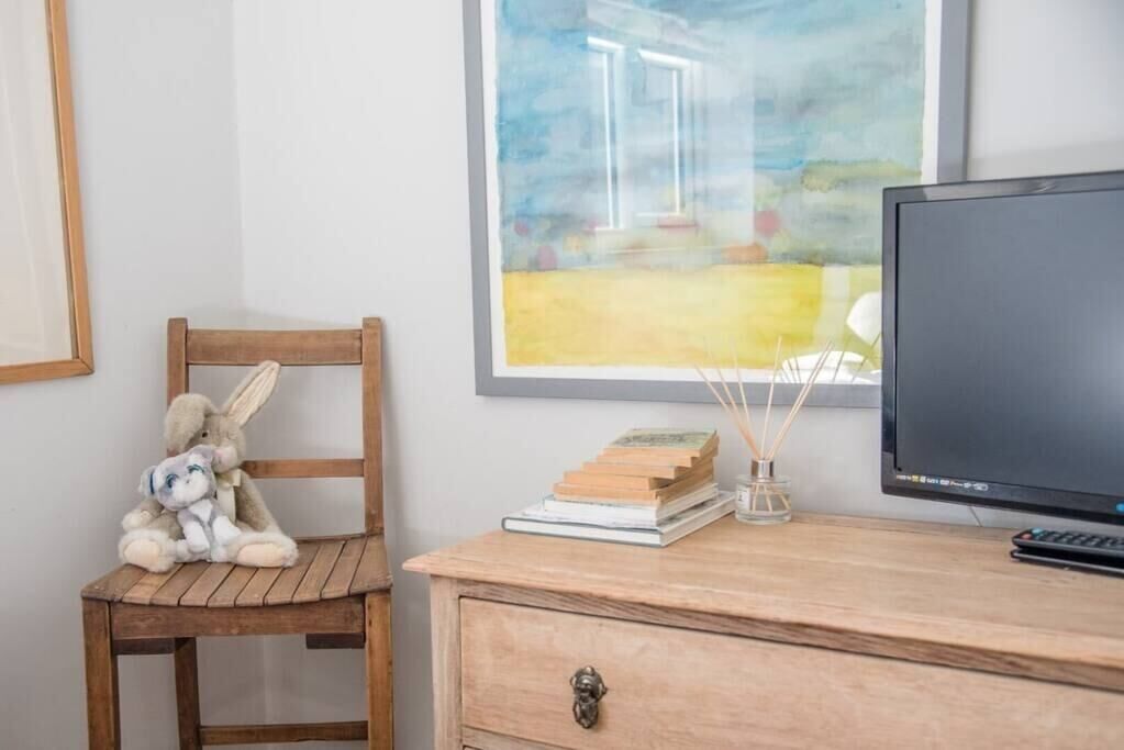 Una silla de madera con un peluche, un escritorio con un monitor de computadora, libros y un pequeño florero con ramas.