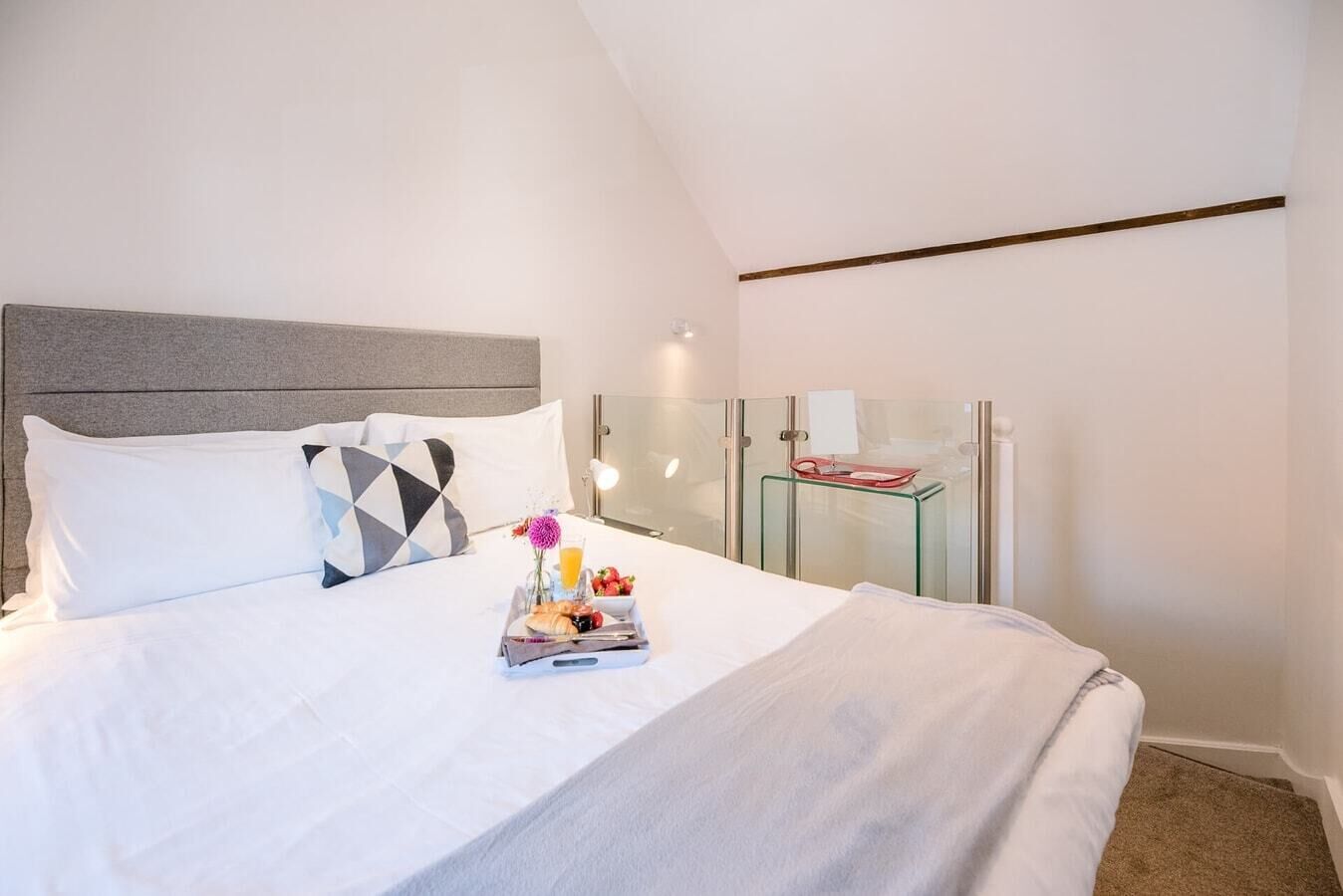 Ein ordentlich bezogenes Bett mit weißen Leinen, ein Tablett mit Frühstücksgebäck und eine moderne Glaswand.