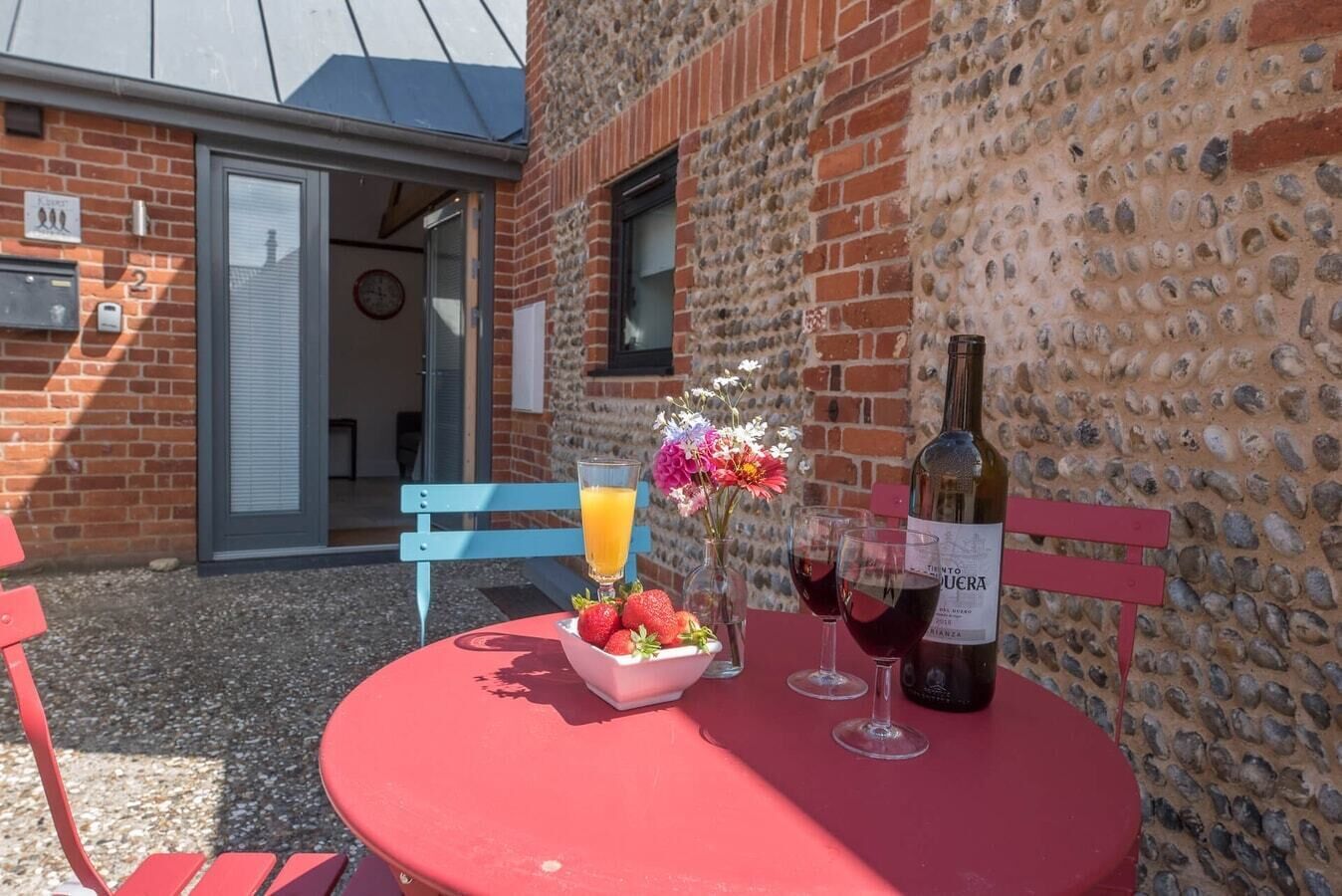 A red table with a bottle of wine, two wine glasses, a bowl of strawberries, and a glass of orange juice.