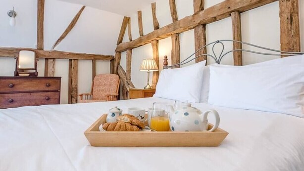 Een slaapkamer met een houten plafond, een bed met witte lakens, een nachtkastje met een lamp en een dienblad met ontbijtartikelen.