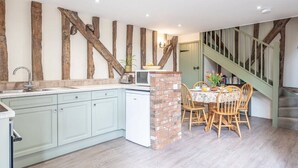 Een keuken met een centraal eiland, een eethoek met tafel en stoelen, en een trap met houten balken.