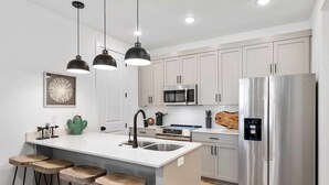Une cuisine moderne avec des meubles blancs, des appareils en acier inoxydable, un îlot central doté de tabourets, des luminaires suspendus et une décoration à thème de cactus.