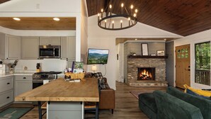 A modern kitchen with a central island, a stone fireplace, and a large window overlooking a scenic view. - Penny Creek Cabin 2 bed 1 bath sleeps 6,Creekside*Hike*Fish*RELAX (Deep Gap)