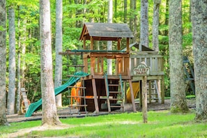 Une structure de jeux en bois, dotée d’un toboggan et d’une zone d’escalade, entourée d’arbres.