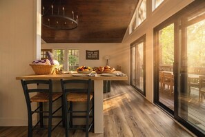 A kitchen with a wooden island, two barstools, a chandelier, and a view of the outdoors through large windows.