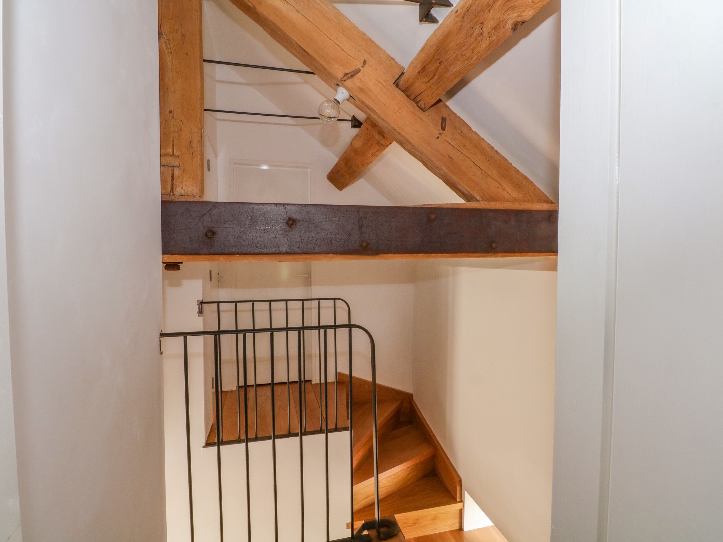 Una escalera de madera con barandillas de metal, una puerta visible en la parte superior y un techo con vigas a la vista.
