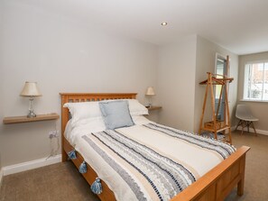 A bedroom with a wooden bed, two bedside tables with lamps, a wooden wardrobe, a chair, and a window with blinds. - Daisy Tree Cottage (Woodhall Spa)