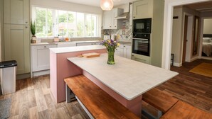 A modern kitchen with a central island, wooden flooring, and a view to the outside. - Daisy Tree Cottage (Woodhall Spa)