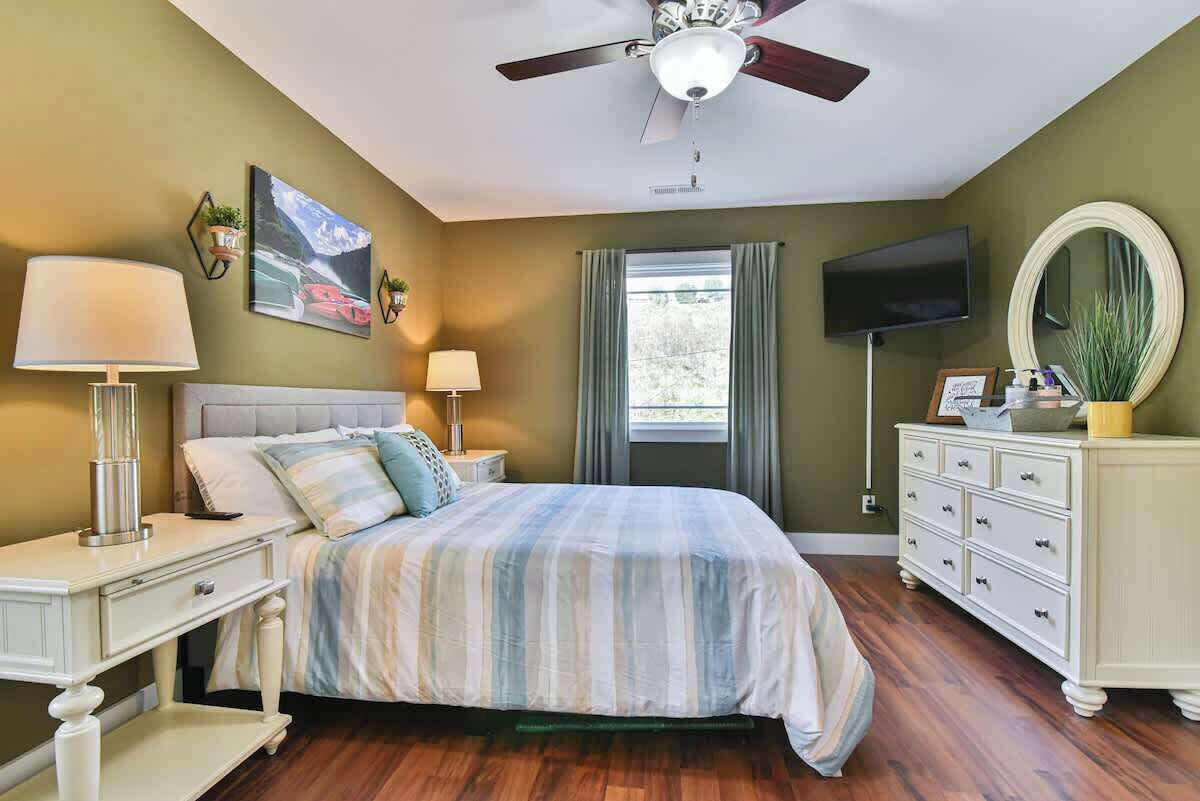 Une chambre à coucher comprenant un lit, des tables de chevet, une commode, un ventilateur de plafond, un téléviseur et une fenêtre avec des rideaux.