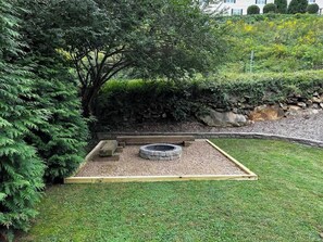 A circular stone fire pit surrounded by a wooden border, set in a grassy area with a stone wall and dense foliage in the background.
