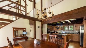 A spacious dining area with a large wooden table and chairs, a chandelier, and a view into a modern kitchen with wooden cabinets and stainless steel appliances.