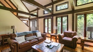 A spacious living room with a high ceiling, exposed wooden beams, and large windows offering a view of the outdoors.