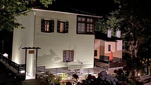 A two-story building with a white facade and dark shutters, surrounded by lush greenery and flowering plants. - DandyVillas Tsagkarada | Sweet Green Apple | Mini Pool (Tsagkarada)