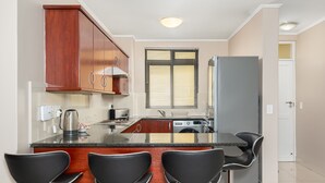 A modern kitchen with a central island, bar stools, wooden cabinets, a refrigerator, and a window with blinds. - Island Club 206 (Cape Town)