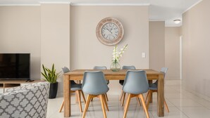 A modern dining area with a wooden table, six grey chairs, a round wall clock, a potted plant, and a television on a stand. - Island Club 206 (Cape Town)
