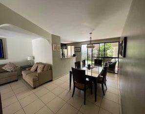 A modern living room with a dining area, a sofa set, and a large window providing natural light.