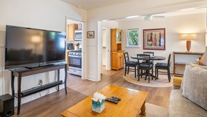 Een woonkamer met een flatscreen televisie, een houten salontafel en een bank. Er is een eethoek met een tafel en stoelen, en een keuken met een oven en kastjes.