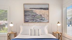 Een slaapkamer met een bed, een schilderij van een strand, twee bedlampen en een raam met zonwering.