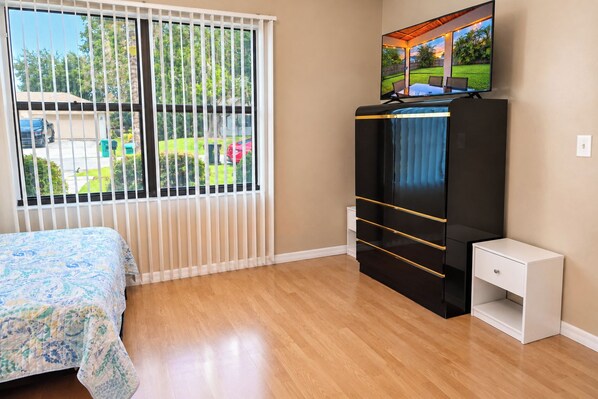 Une chambre à coucher avec un lit, un téléviseur posé sur un meuble, une fenêtre avec des stores et une vue sur l’extérieur.