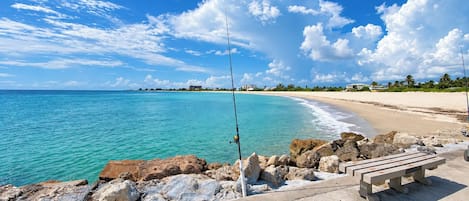 Uma praia com águas turquesa cristalinas, costão rochoso e calçada de concreto com um banco.